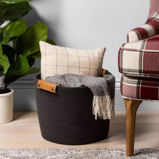 The Brightroom™ gray coiled rope basket in a cozy room setting, filled with a plush throw blanket, next to a plant and a wooden-legged chair.