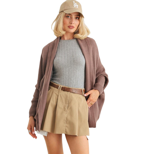 A model in a cozy knit cardigan with batwing sleeves and ribbed texture, styled over a grey top and khaki skirt, accessorized with a beige baseball cap, showcasing a casual yet stylish look.