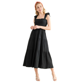 Woman in a black smocked sundress with ruffled straps and a tiered skirt. The dress has a fitted bodice and a flared, ankle-length hem. The model is standing with one hand resting lightly on her other hand, showcasing the dress's elegant and comfortable style.