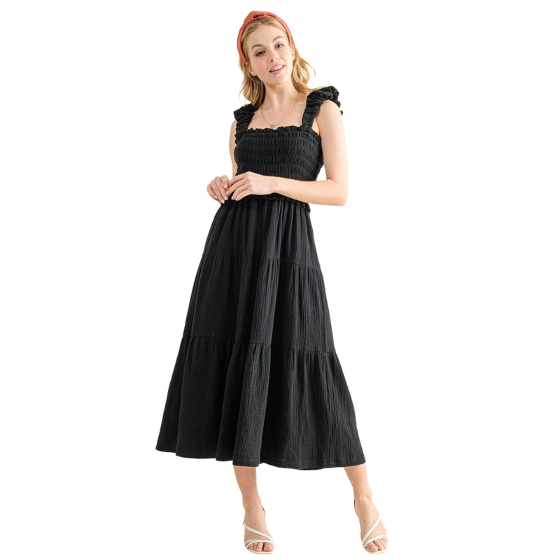 Load image into Gallery viewer, Woman in a black smocked sundress with ruffled straps and a tiered skirt. The dress has a fitted bodice and a flared, ankle-length hem. The model is standing with one hand resting lightly on her other hand, showcasing the dress&#39;s elegant and comfortable style.
