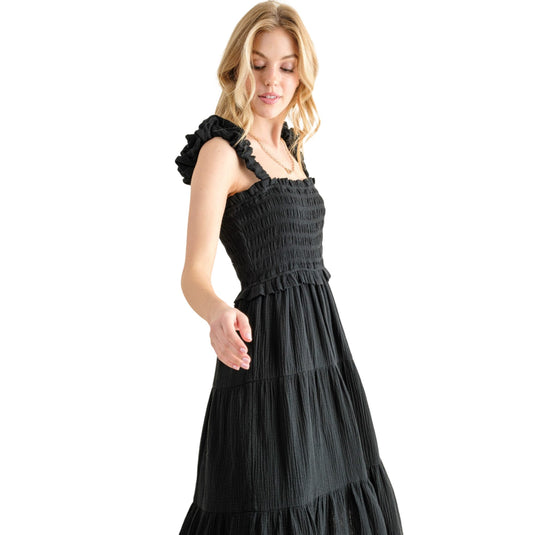 Woman wearing a black smocked sundress with ruffled straps and a tiered skirt. The dress features a fitted bodice and a flowing, ankle-length hem, perfect for a casual summer day. The model is shown looking down, showcasing the dress's elegant and relaxed fit.