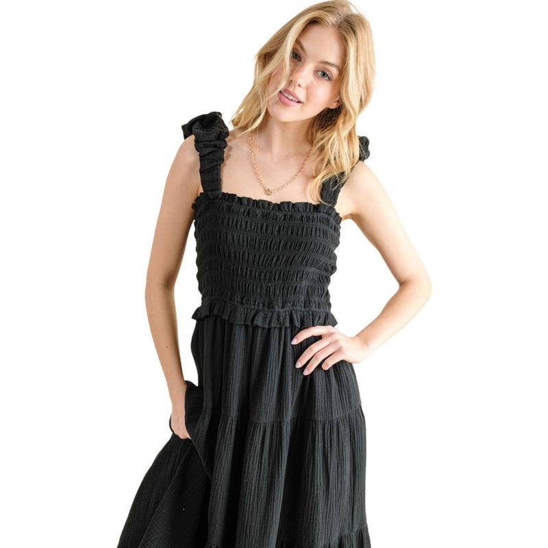 Load image into Gallery viewer, Model posing in a black smocked sundress with ruffled straps and a tiered skirt. The dress has a fitted bodice and a flared, ankle-length hem. The model has one hand on her hip, highlighting the dress&#39;s flattering silhouette and comfortable fit.
