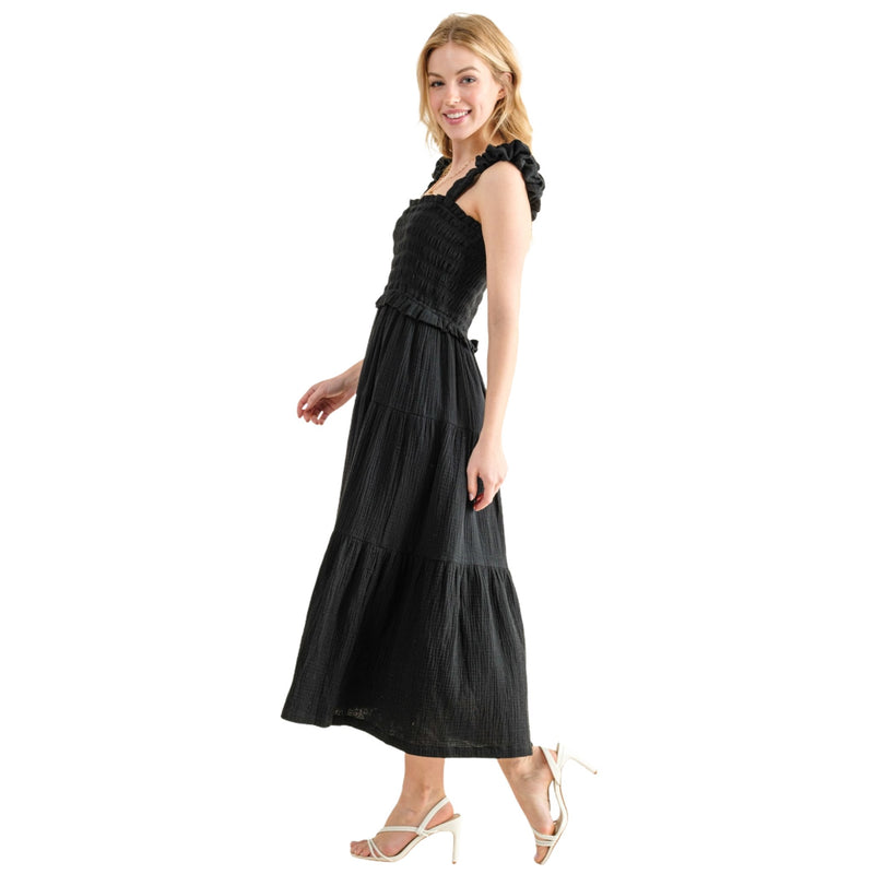 Load image into Gallery viewer, Side view of a woman wearing a black smocked sundress with ruffled straps and a tiered skirt. The dress features a fitted bodice and a flowing, ankle-length hem, perfect for casual outings. The model is walking, showing the dress&#39;s movement and flow.
