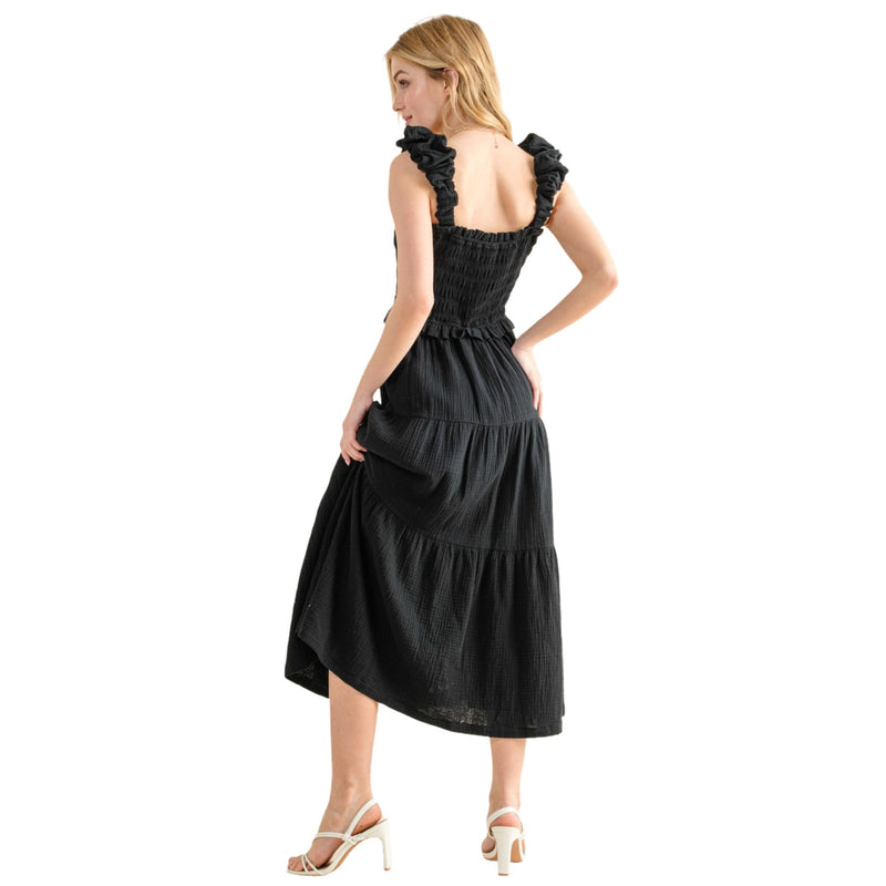 Load image into Gallery viewer, Back view of a woman wearing a black smocked sundress with ruffled straps and a tiered skirt. The dress features a fitted bodice and a flowing, ankle-length hem, ideal for casual summer days. The model&#39;s posture highlights the dress&#39;s back design and relaxed fit.
