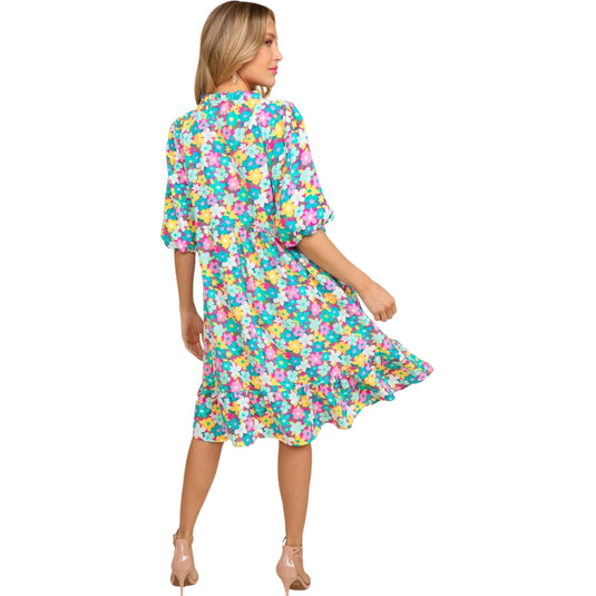 Back view of a woman wearing a vibrant bubble sleeve floral midi dress with ruffles in a mint and pink color scheme. She is looking to the side, with one hand gently holding the hem of her dress.