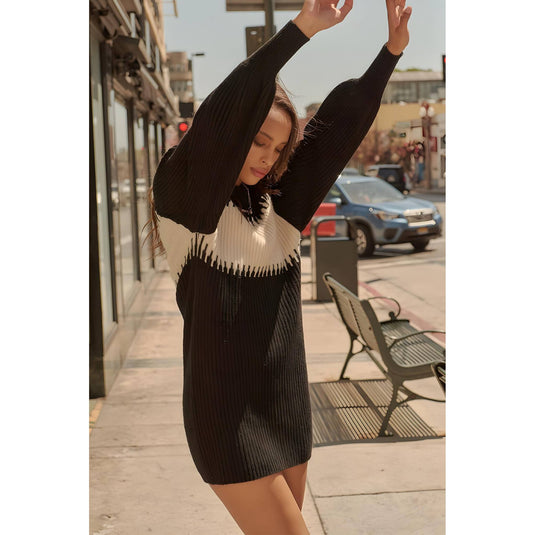 Stylish young lady stretches gracefully on a sunny street corner, dressed in a chic black sweater dress with distinctive white colorblock design, complemented by modern, chunky grey boots.