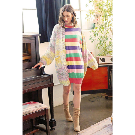 A model stands contemplatively beside an old piano in a warmly lit room, wearing a Coral Combo Striped Sweater Dress paired with a chunky pastel cardigan. The dress features vivid horizontal stripes in coral, green, yellow, navy, and white, complemented by the cardigan’s soft, multicolored yarn. She completes her outfit with mid-calf suede boots, perfect for a cozy, artistic vibe.