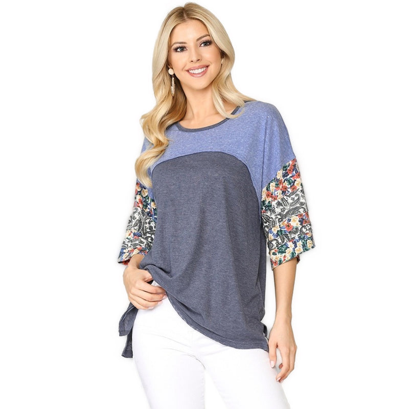 Load image into Gallery viewer, A woman wearing a stylish top with a denim mix design. The top features a blue and grey color block pattern with floral dolman sleeves, paired with white pants. She is smiling and has long blonde hair.
