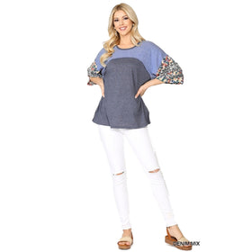 Full-body view of a woman dressed in a denim mix floral dolman top and white ripped jeans. The top has a blue and grey color block design with floral patterned sleeves. She is standing with her hands on her hips and smiling.