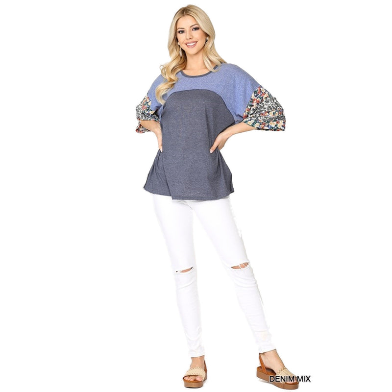 Load image into Gallery viewer, Full-body view of a woman dressed in a denim mix floral dolman top and white ripped jeans. The top has a blue and grey color block design with floral patterned sleeves. She is standing with her hands on her hips and smiling.
