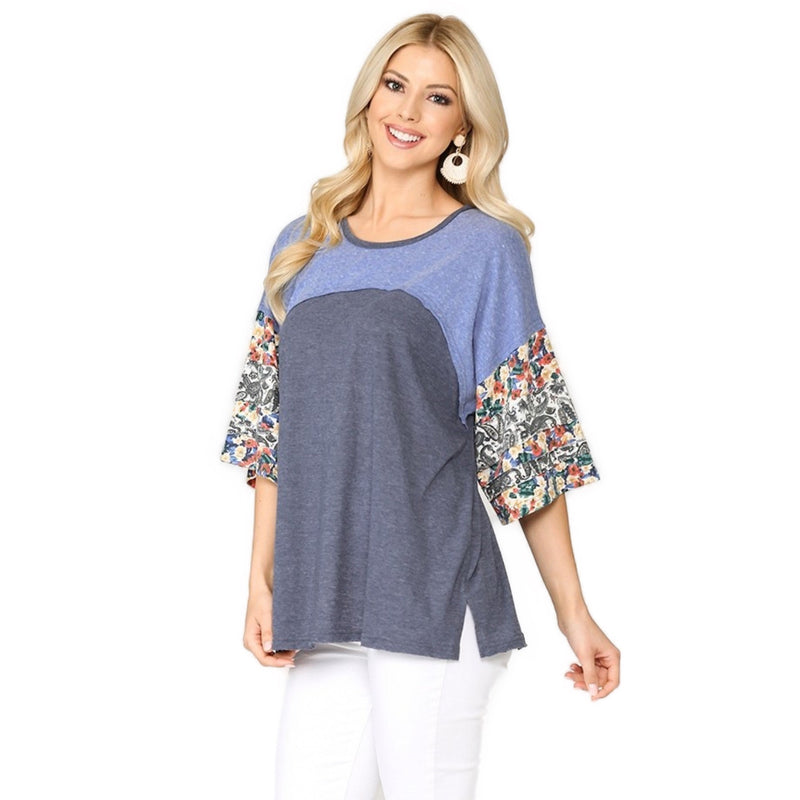 Load image into Gallery viewer, A woman smiling while wearing a denim mix floral dolman top. The top has a blue and grey color block pattern with colorful floral sleeves. She is paired with white pants and large round earrings.
