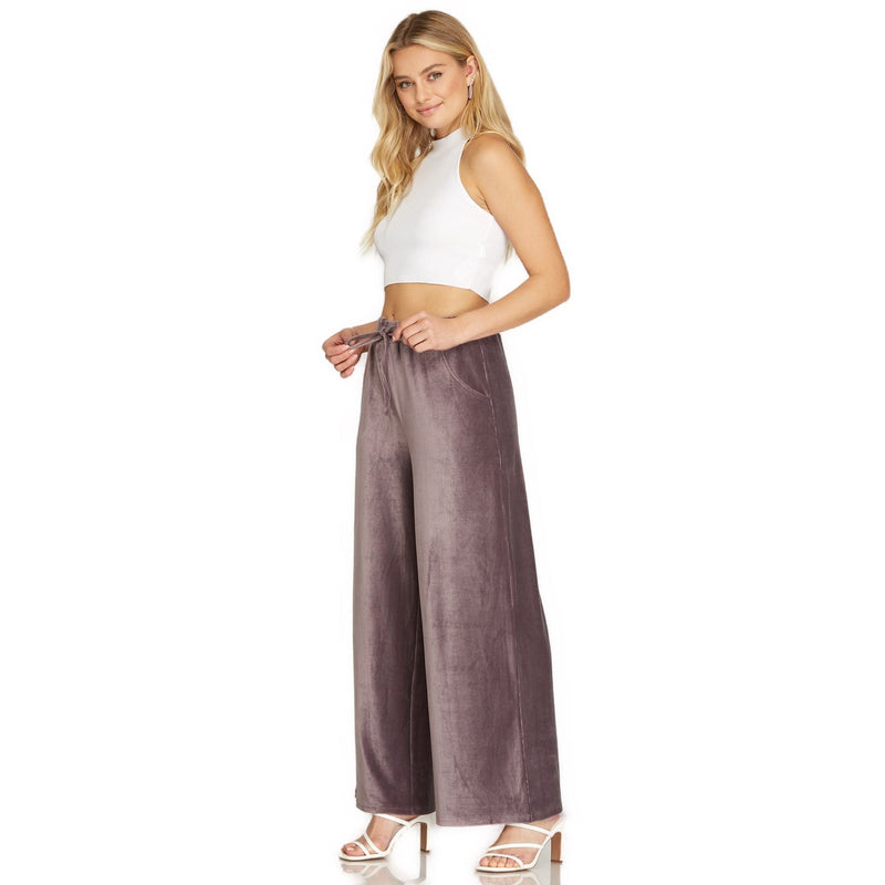 Load image into Gallery viewer, Model turned slightly, giving a three-quarter view of the velvet wide-leg pants in mauve with elastic waistband and pockets, complemented by a white crop top.
