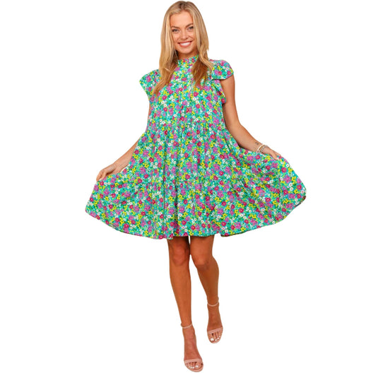 Full-body view of a woman wearing a bright floral print dress with a high neckline and short ruffled sleeves. The dress has a vibrant mix of green, pink, and blue flowers, with a loose, above-the-knee fit. The model is holding out the sides of the dress, showing off its flowy and playful design.