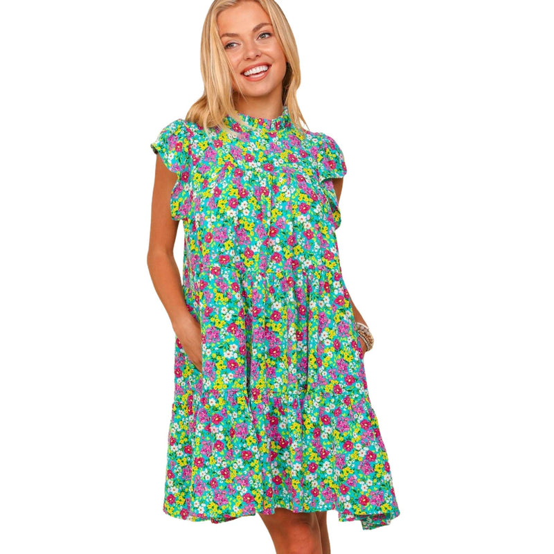 Load image into Gallery viewer, Front view of a woman wearing a bright floral print dress with a high neckline and short ruffled sleeves. The dress has a vibrant mix of green, pink, and blue flowers, with a relaxed, above-the-knee fit. The model is standing with her hands in the dress&#39;s pockets, exuding a casual and cheerful vibe.
