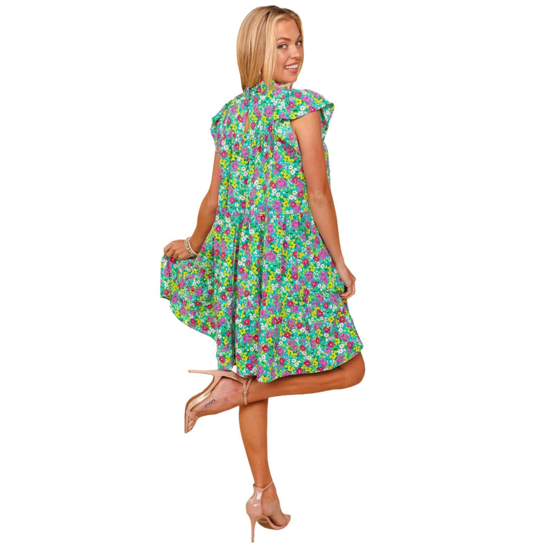 Load image into Gallery viewer, Back view of a woman wearing a bright floral print dress with a high neckline and short ruffled sleeves. The dress features a vibrant mix of green, pink, and blue flowers, with a keyhole detail at the back and a loose, above-the-knee fit. The model is smiling and lifting one foot slightly, highlighting the dress&#39;s playful and comfortable style.
