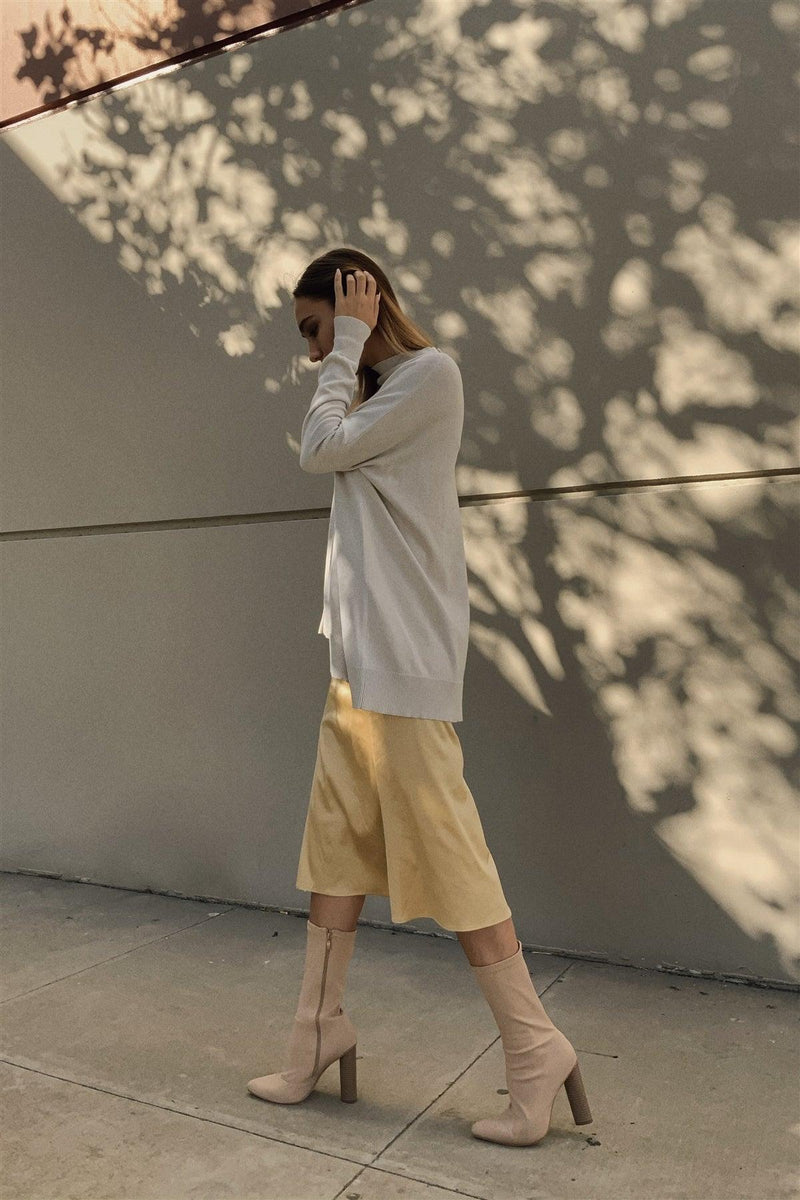Load image into Gallery viewer, A woman in a relaxed, contemplative pose wearing a golden satin midi skirt paired with a long-sleeve white top and beige knee-high boots, set against a backdrop with a play of light and shadow.

