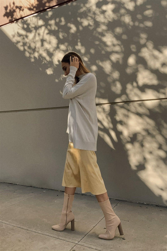 A woman in a relaxed, contemplative pose wearing a golden satin midi skirt paired with a long-sleeve white top and beige knee-high boots, set against a backdrop with a play of light and shadow.