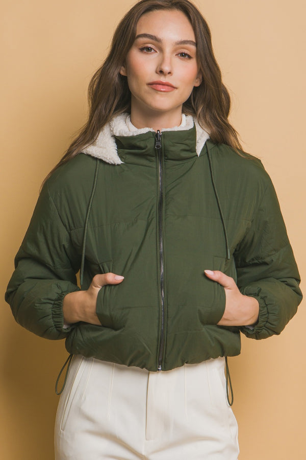 Image of a model wearing a green cropped Sherpa reversible jacket, facing forward and showcasing the zipper and drawstring hood.