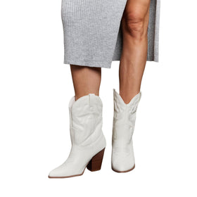 A close-up view of a model wearing white mid-calf cowboy boots with block heels, paired with a ribbed gray midi skirt. The boots feature a classic Western design with detailed stitching and a pointed toe.