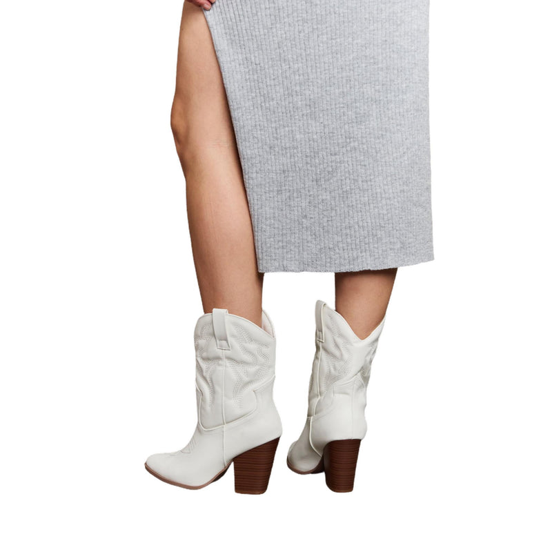 Load image into Gallery viewer, The back view of the white cowboy boots, highlighting the intricate stitching and the block heel. The boots are paired with a ribbed gray skirt with a slit, showcasing the elegant yet rugged look of the footwear.
