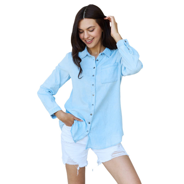 Woman standing in a light blue chambray denim button-down long sleeve shirt paired with white distressed shorts, smiling and holding her hair.