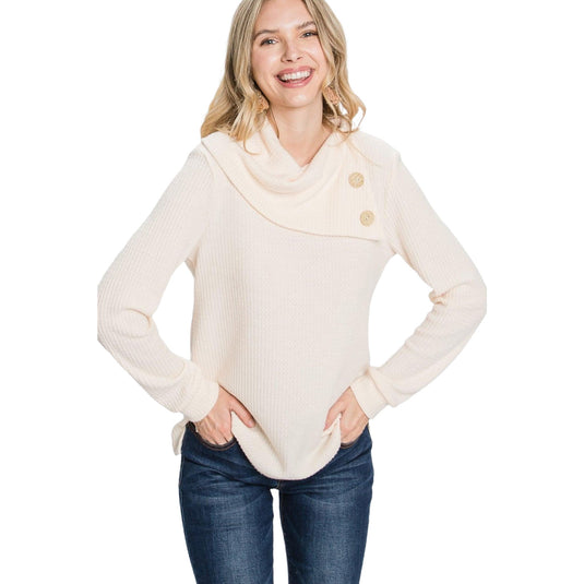 A cheerful woman in an off-white mock neck sweater with stylish button details on the shoulder, hands casually placed in the front pockets of her blue jeans.