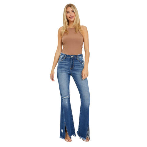 Full-body front view of a woman wearing plus-size high-rise flare jeans with a front slit and frayed hem, paired with a sleeveless brown top. These trendy jeans offer a flattering fit with a retro-inspired silhouette and modern distressed detailing.