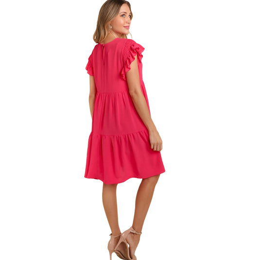  Back view of a woman wearing a bright pink, ruffle cap sleeve, crew neck dress with a tiered skirt and pockets. She is standing with her arms relaxed by her side, looking over her shoulder.