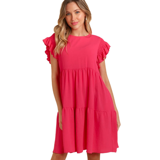 Front view of a woman wearing a bright pink, ruffle cap sleeve, crew neck dress with a tiered skirt and pockets. She is standing with her hands relaxed at her sides and a slight smile on her face.