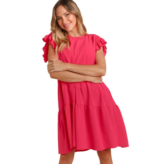 Front view of a woman wearing a bright pink, ruffle cap sleeve, crew neck dress with a tiered skirt and pockets. She is standing with her arms crossed and a big smile on her face.