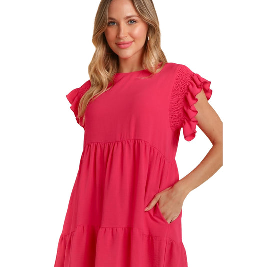 Close-up front view of a woman wearing a bright pink, ruffle cap sleeve, crew neck dress with a tiered skirt and pockets. She has one hand in her pocket and is smiling softly.