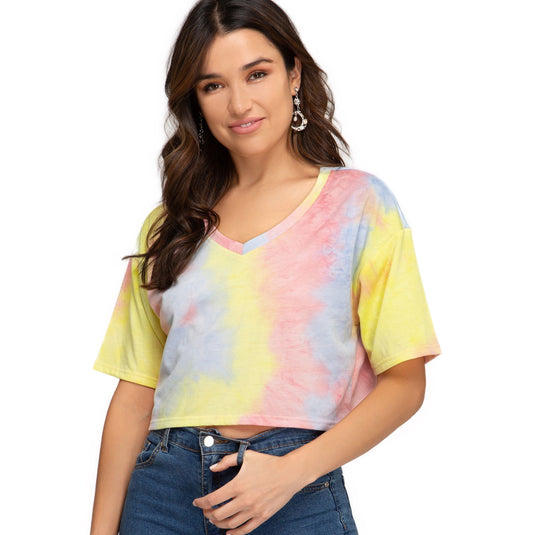 A woman models a Short Sleeve V Neck Terry Crop Top with a vibrant yellow and pink tie-dye pattern, paired with classic blue jeans. The crop top is designed for a relaxed fit and is available in sizes S, M, L.