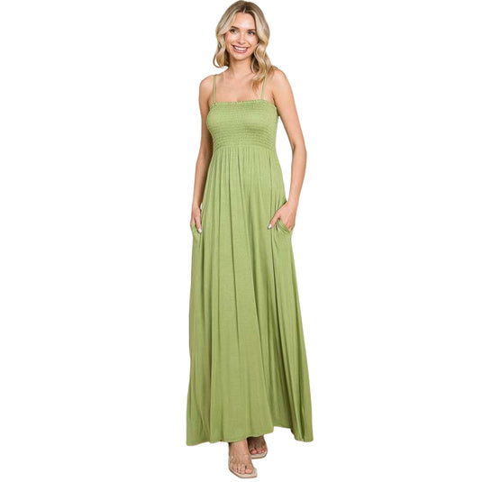 Front view of a woman wearing a light green maxi dress with a smocked bodice and thin straps. The dress features a flowing skirt and pockets, offering both style and practicality. The model is smiling and exudes a relaxed, comfortable look.