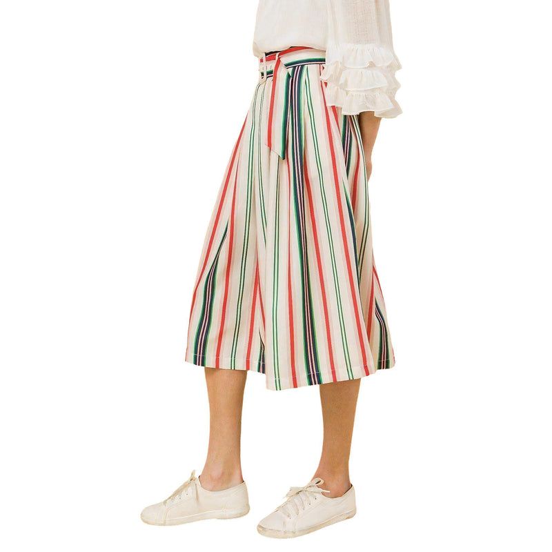 Load image into Gallery viewer, Side view of a model showcasing a striped pleated midi skirt with pockets, paired with a white blouse and white sneakers, highlighting the skirt&#39;s flow and mid-calf length.
