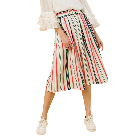 A model in profile view wearing a pleated midi skirt with vertical stripes in shades of red, green, and beige, complemented by a white blouse with ruffle details and casual white sneakers.