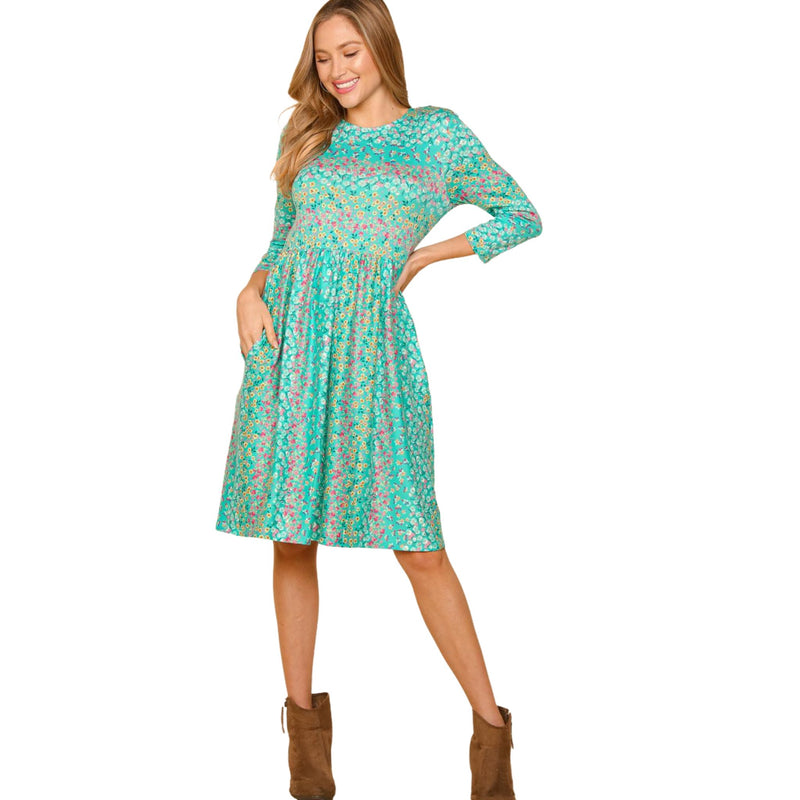 Load image into Gallery viewer, Woman in a teal floral dress with 3/4 sleeves, posing with one hand on her hip and the other in the dress pocket.
