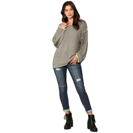 A full-length image of a woman standing confidently in a grey two-tone sweater with contrasting piping, complemented by distressed blue jeans and black ankle boots.