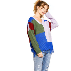 A model poses with her hand on her head, wearing a vibrant, oversized color-block sweater with a fringed hem and distressed blue jeans.