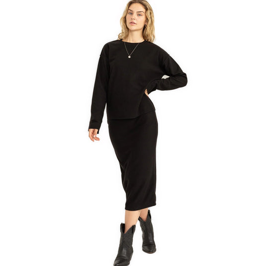 A woman stands in a relaxed pose wearing a black high-waisted midi skirt paired with a matching sweater and black cowboy boots, exuding a casual yet chic autumn style.