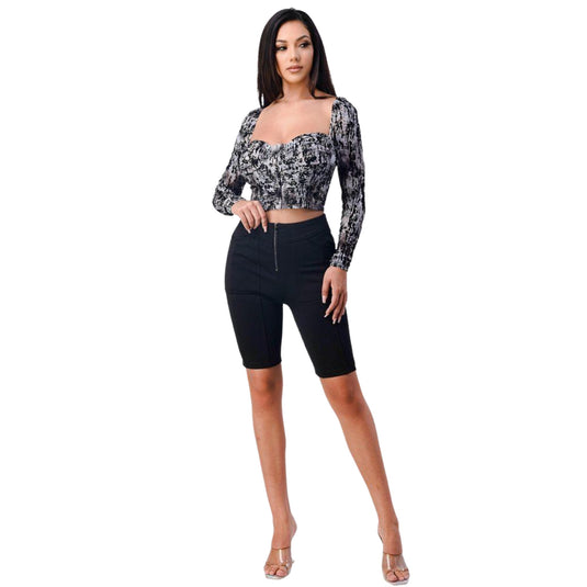 The image captures a stylish woman sporting black zip-up biker shorts complemented by a trendy, long-sleeve black and white patterned crop top, complete with a square neckline. She stands confidently, showing off a versatile outfit that merges comfort with high street style.